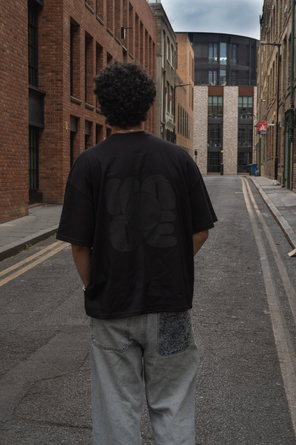 Person wearing a black t-shirt and light gray pants standing on a city street with brick buildings.